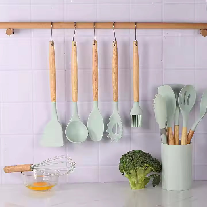 Set of green kitchen utensils with wooden handles hanging on a rack against a tiled wall.
